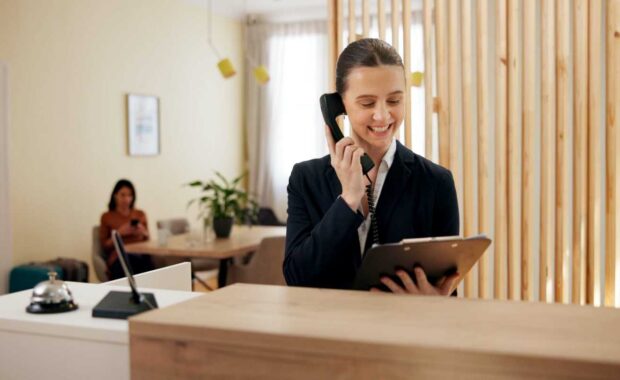 Smiling Female Concierge with Clipboard Talking Over Phone at Her Desk