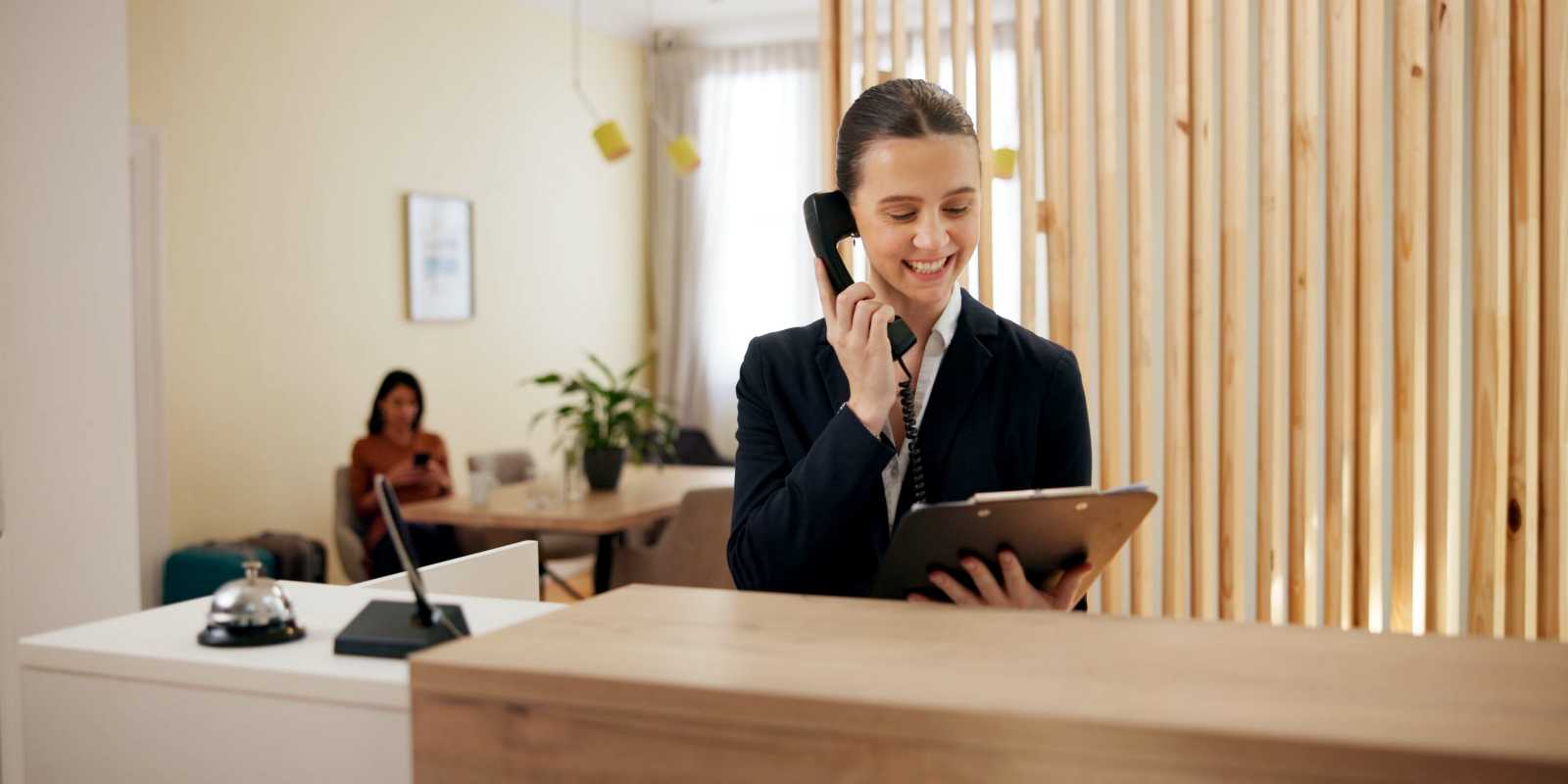 Smiling Female Concierge with Clipboard Talking Over Phone at Her Desk